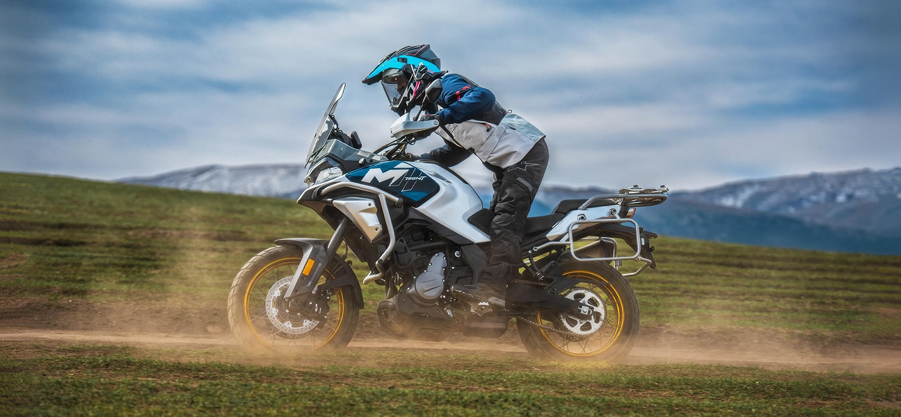 CFmotoMartinez - moto CFMOTO 700MT ADV circulando sobre tierra con conductor de pie y fondo de montaña